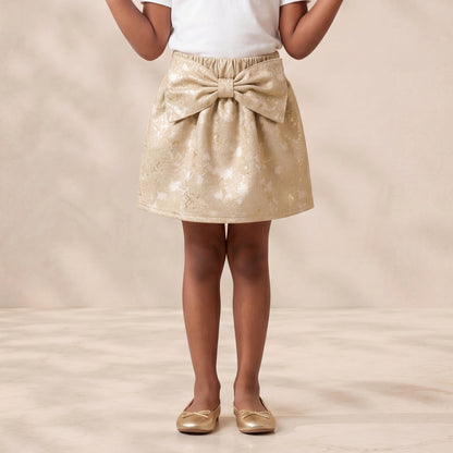 A young girl stands smiling with her hands raised, wearing a white short-sleeve t-shirt and a shiny gold sequined mini skirt with a large bow at the waist, paired with gold flats, against a warm beige textured studio backdrop.