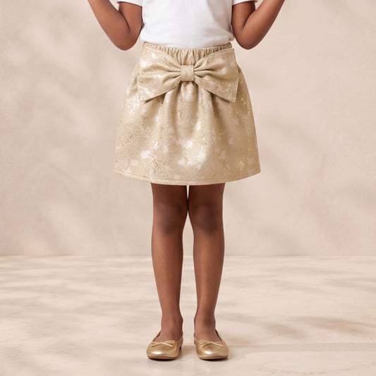 A young girl stands smiling with her hands raised, wearing a white short-sleeve t-shirt and a shiny gold sequined mini skirt with a large bow at the waist, paired with gold flats, against a warm beige textured studio backdrop.