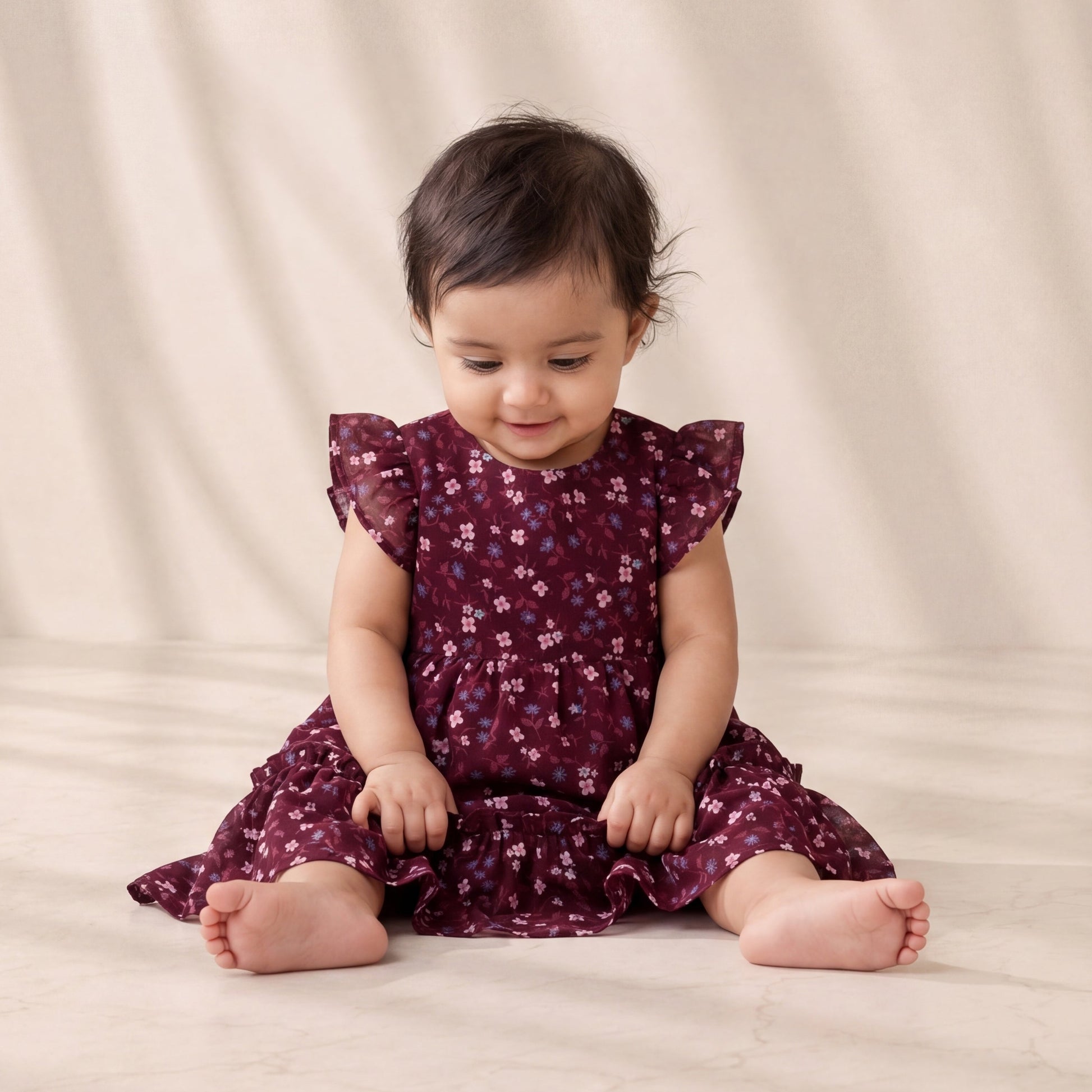 Baby in a purple floral dress sitting on a beige surface with a beige curtain background