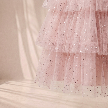 A close-up of a light pink tiered tulle skirt with scattered silver sequins, set against a warm beige background with soft shadows.