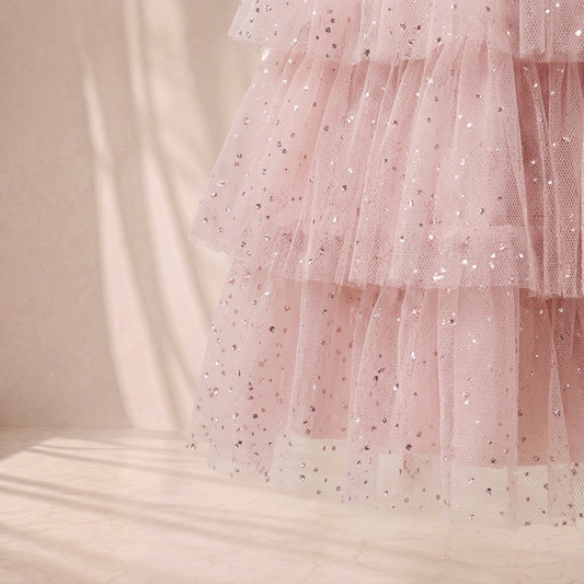 A close-up of a light pink tiered tulle skirt with scattered silver sequins, set against a warm beige background with soft shadows.