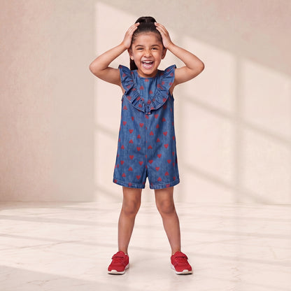 Young girl wearing a blue denim dress with red heart patterns and red shoes, standing against a plain background.