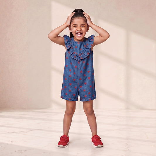 Young girl wearing a blue denim dress with red heart patterns and red shoes, standing against a plain background.