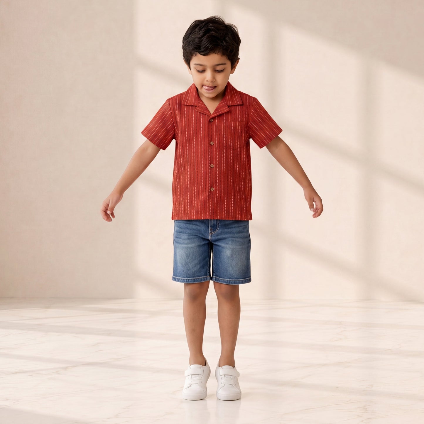 Child wearing a red striped shirt and blue shorts standing on a light wooden floor with a neutral background