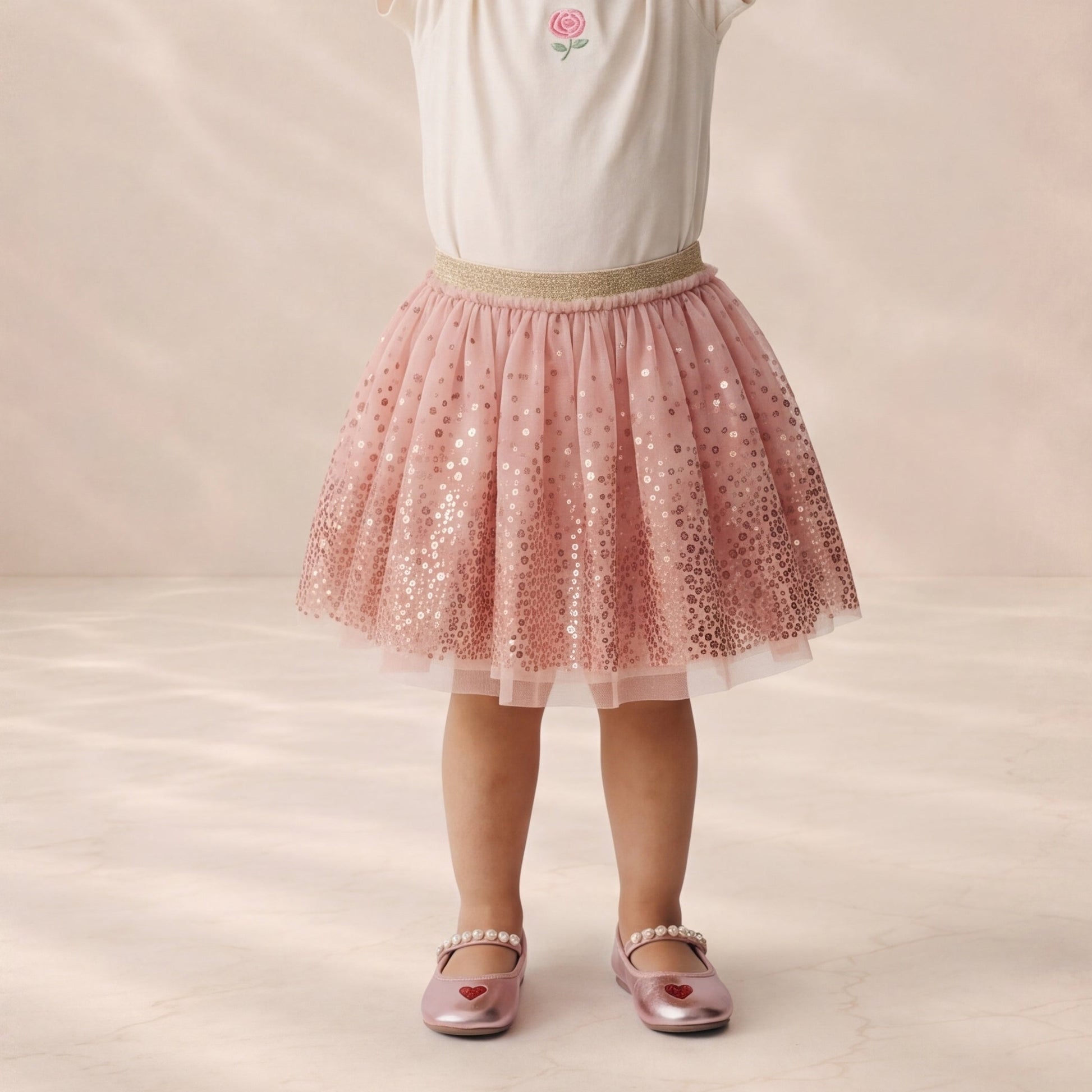 A child wearing a pink sequined tulle tutu skirt with a beige waistband, paired with a white top and metallic pink shoes, standing against a soft beige studio backdrop.