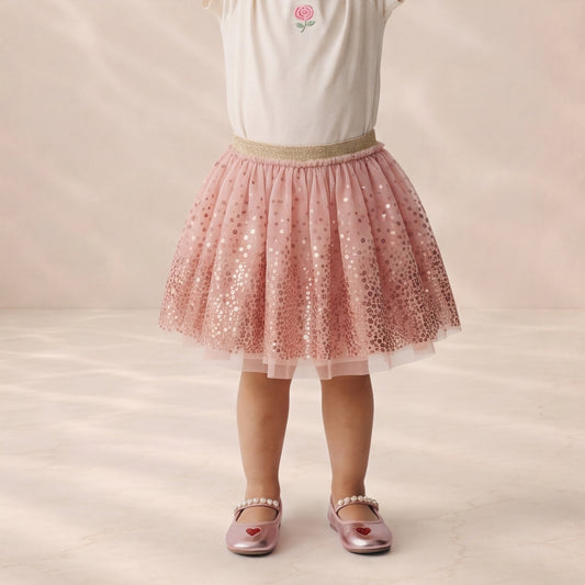A child wearing a pink sequined tulle tutu skirt with a beige waistband, paired with a white top and metallic pink shoes, standing against a soft beige studio backdrop.
